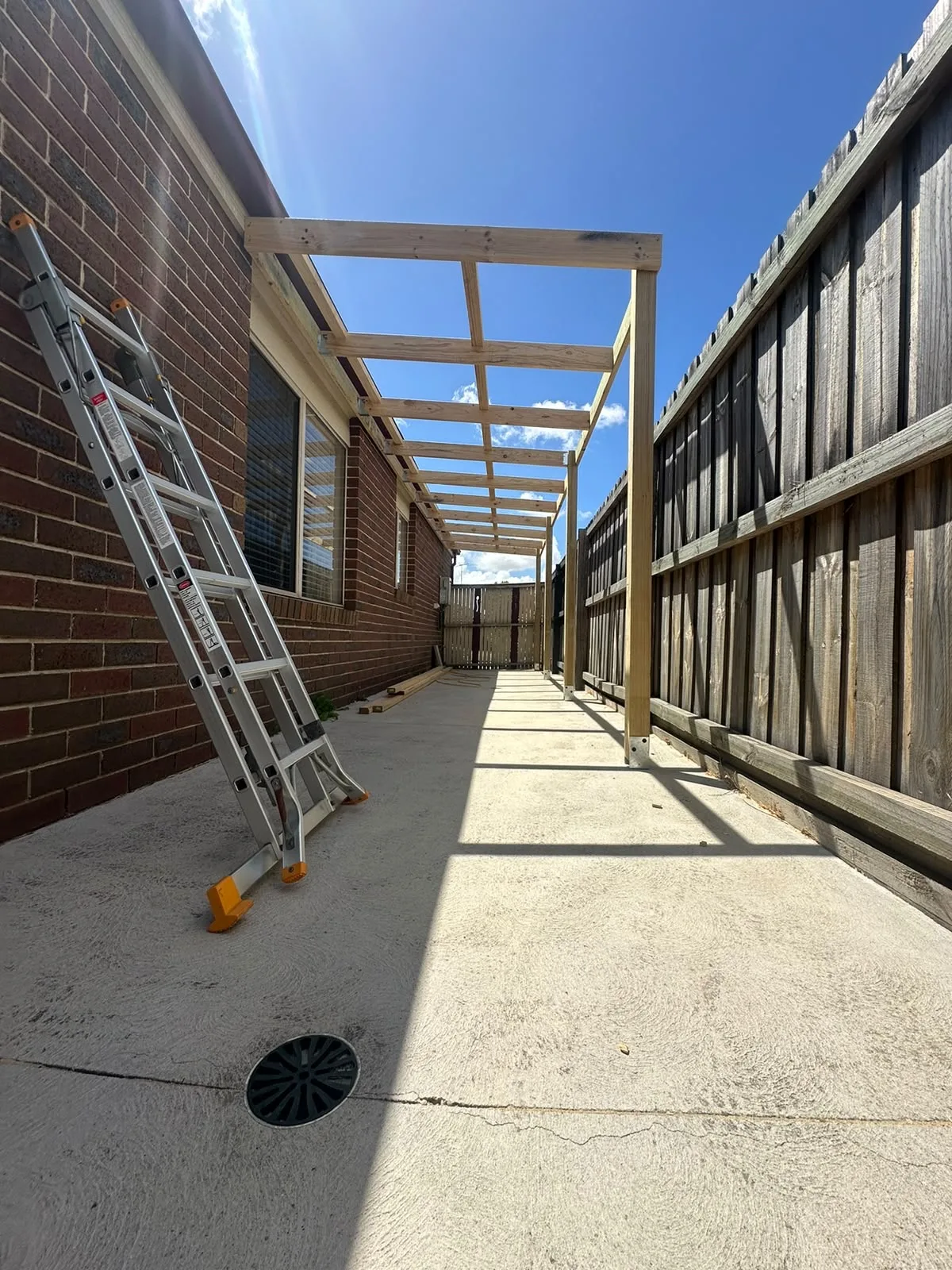 Treated pine pergola frame erected over a concrete side path between a brick house and paling fence