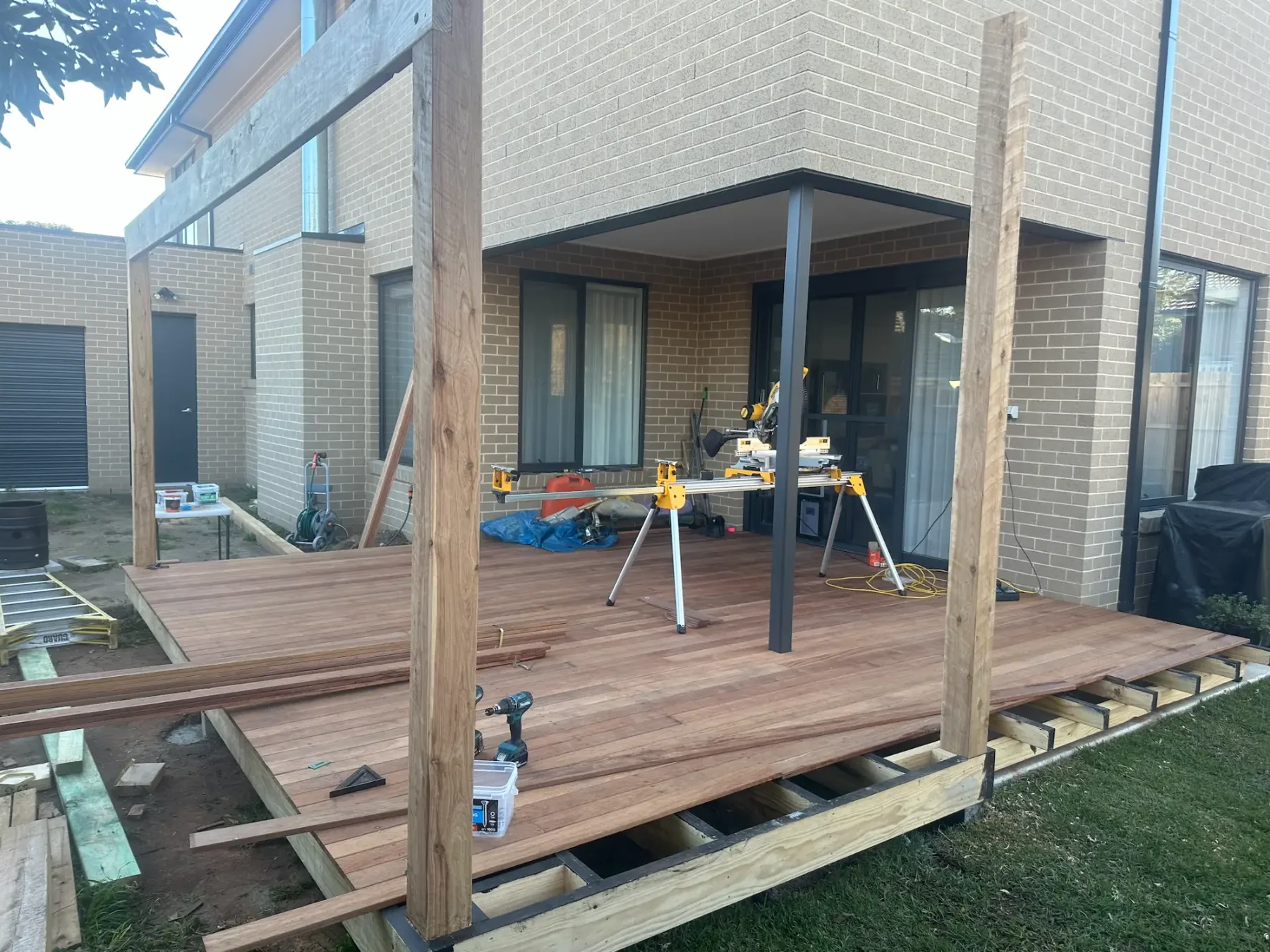 Hardwood deck being laid under a brick alfresco with tall pergola posts standing and a drop saw set up on site
