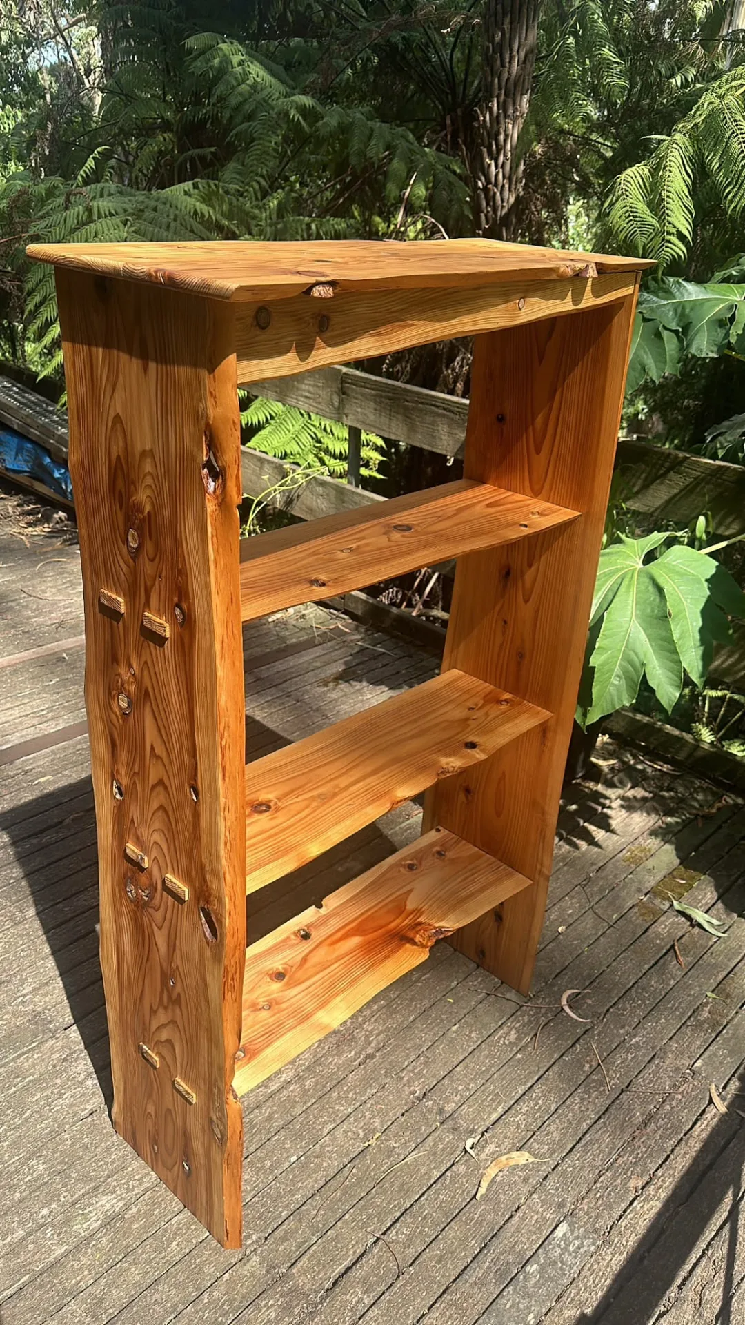Handmade cypress bookshelf with live edges and exposed timber plugs, standing on a fern-lined deck