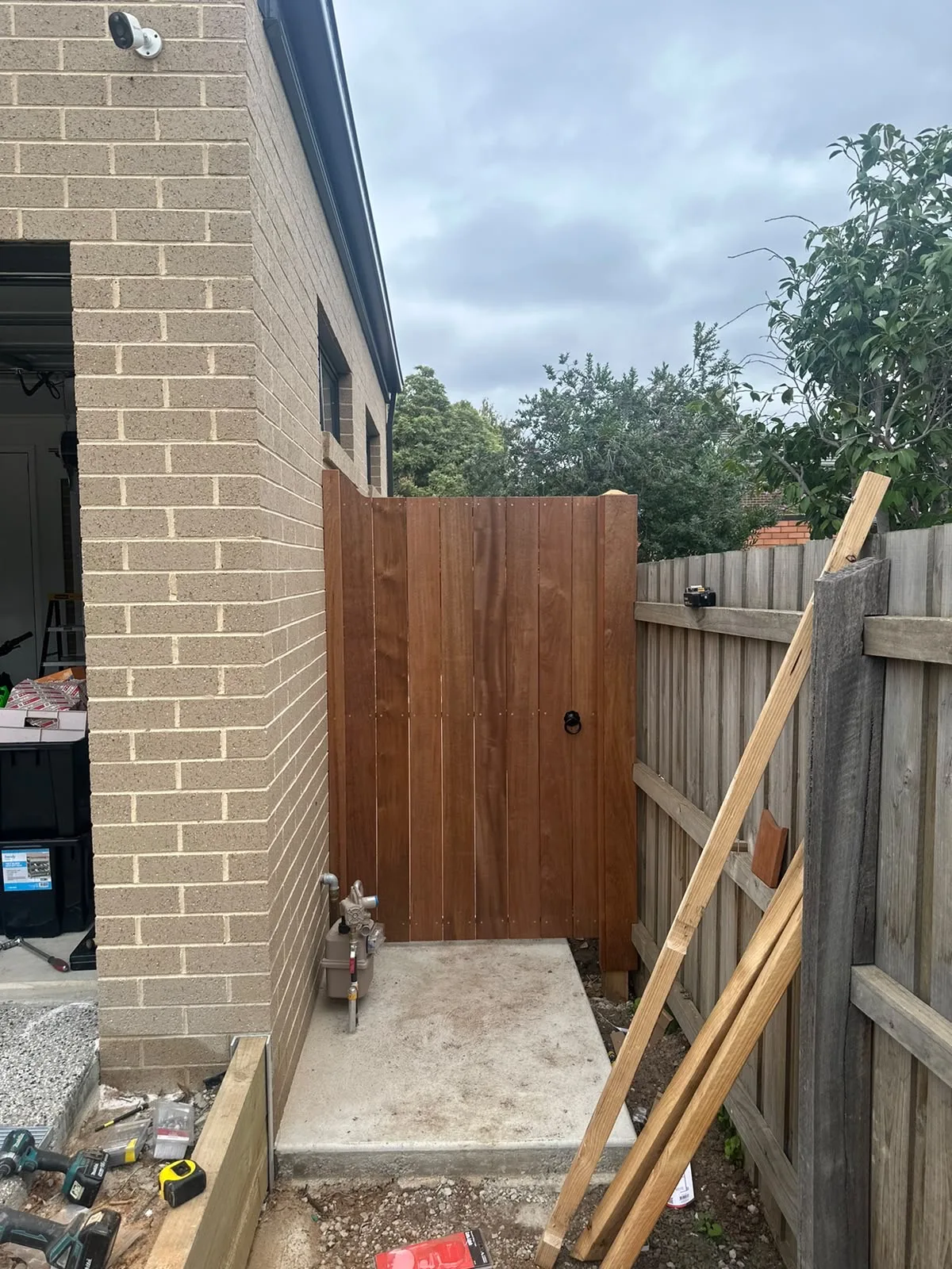 Tall hardwood side gate hung between a brick wall and paling fence on a concrete pad